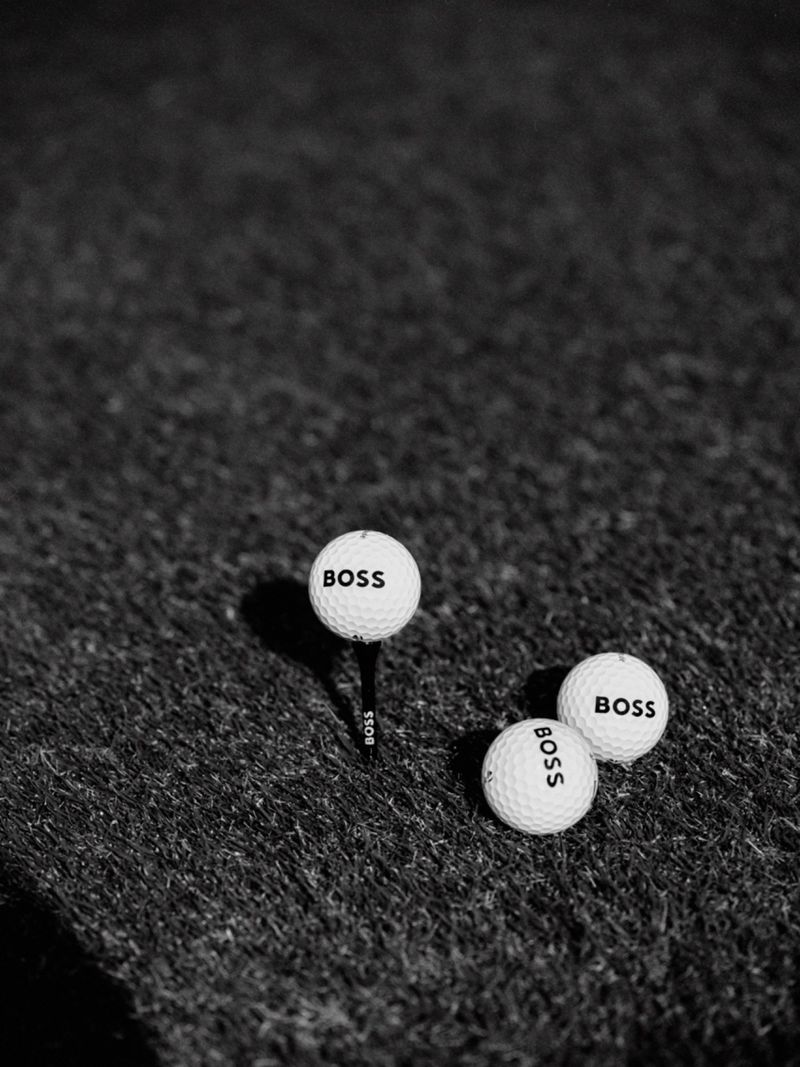 Black and white shot of three golf balls on a putting green, one ball is on a tee, all three golf balls have 'BOSS' printed on them.