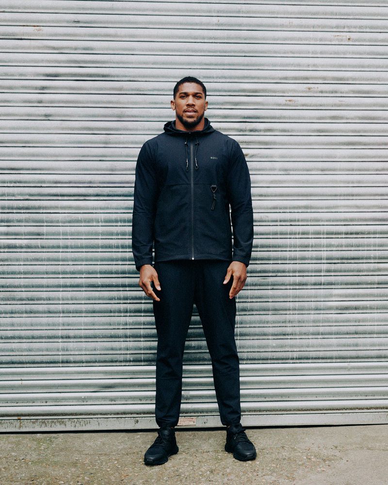 Full-length portrait of a man in dark athletic apparel standing against a corrugated metal background, creating a simple and focused composition.