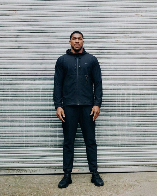 Full-length portrait of a man in dark athletic apparel standing against a corrugated metal background, creating a simple and focused composition.