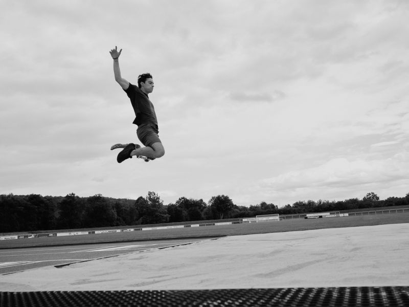 Monochrome shot of a male figure leaping against a cloudy sky, capturing movement and energy with trees and a track in the background.