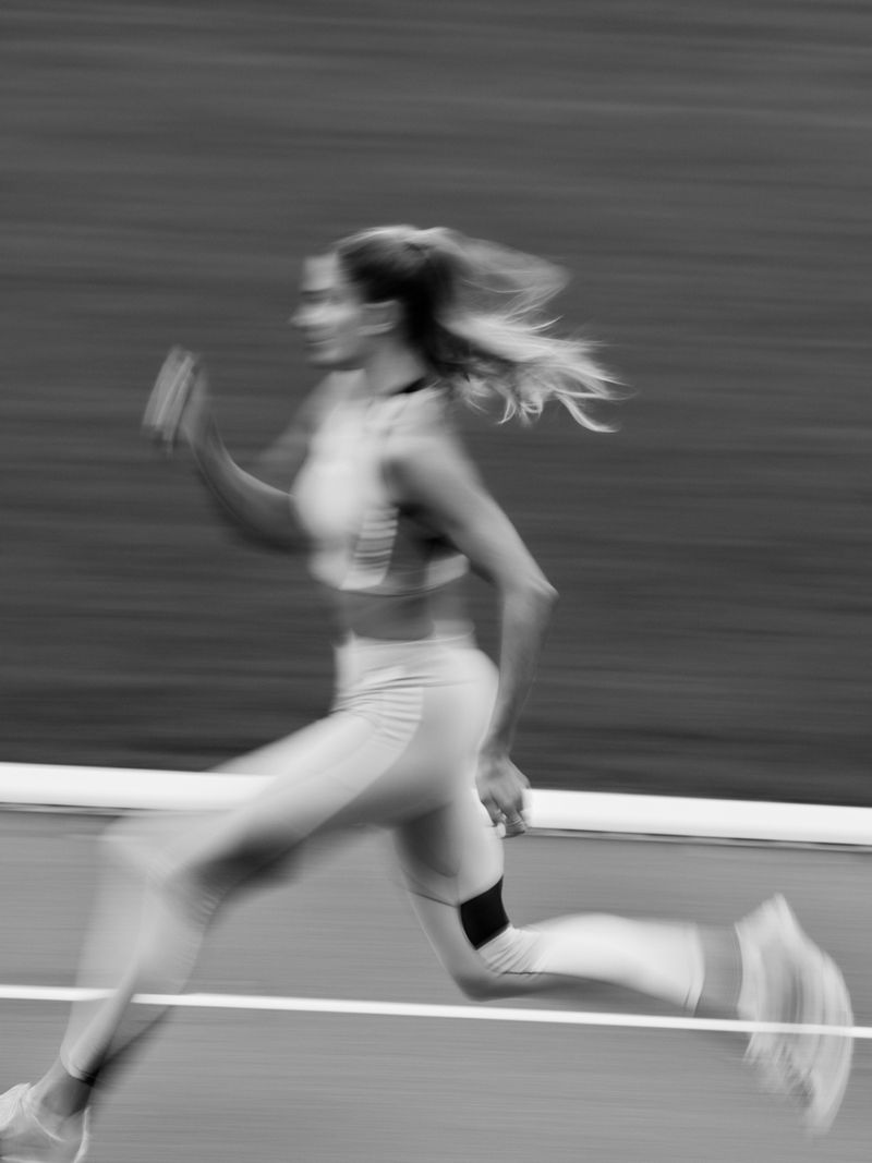 Monochrome long exposure of a woman running on a track, capturing motion blur and a sense of speed in an outdoor setting.