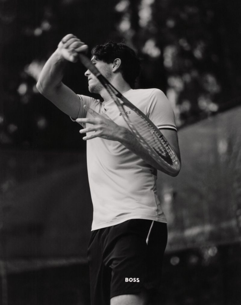 Monochrome shot of a male tennis player with a racket raised, poised to strike a ball, set against a blurred outdoor backdrop.