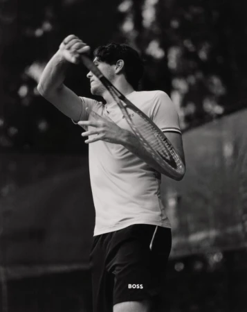 Monochrome shot of a male tennis player with a racket raised, poised to strike a ball, set against a blurred outdoor backdrop.