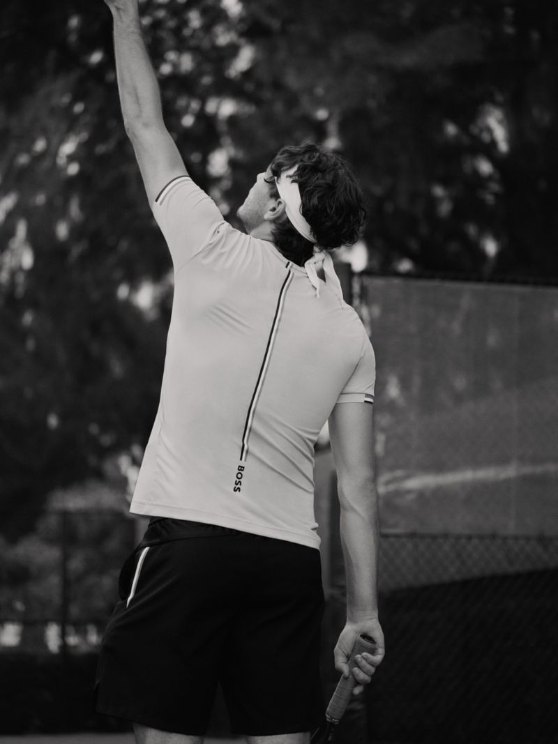Black and white shot of an adult male tennis player serving on an outdoor court, wearing sportswear and headband, back view.