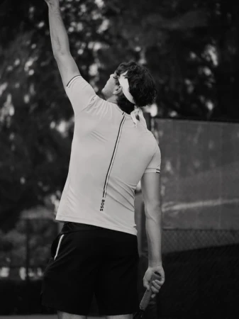 Black and white shot of an adult male tennis player serving on an outdoor court, wearing sportswear and headband, back view.
