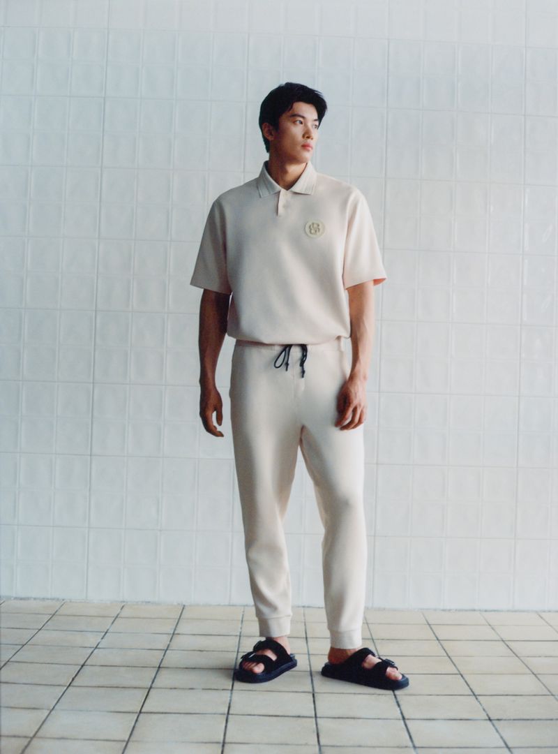 Studio shot of a young adult male model wearing a beige polo shirt and sweatpants, posing on a tiled floor in neutral setting.