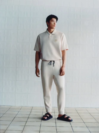 Studio shot of a young adult male model wearing a beige polo shirt and sweatpants, posing on a tiled floor in neutral setting.