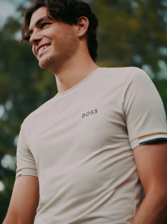 Taylor Fritz smiling while wearing a cream-colored BOSS athletic t-shirt outdoors against a blurred green park background.