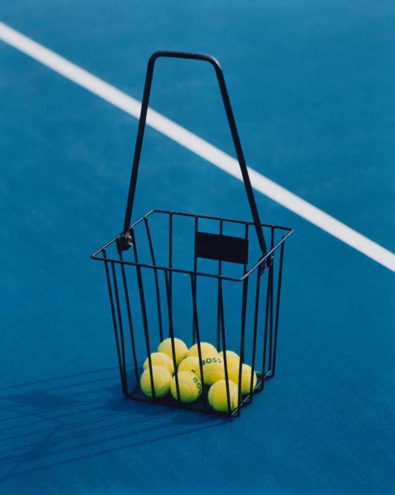 Tennis balls in a black wire basket on a blue tennis court with a white line in the background. Sport and recreation visual.
