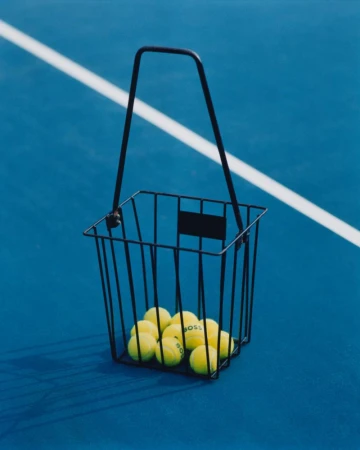 Tennis balls in a black wire basket on a blue tennis court with a white line in the background. Sport and recreation visual.