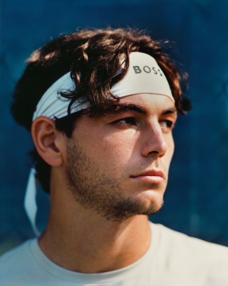 Close-up portrait of an adult male with brown hair wearing a white headband with the text BOSS, and a white t-shirt on a blue background.