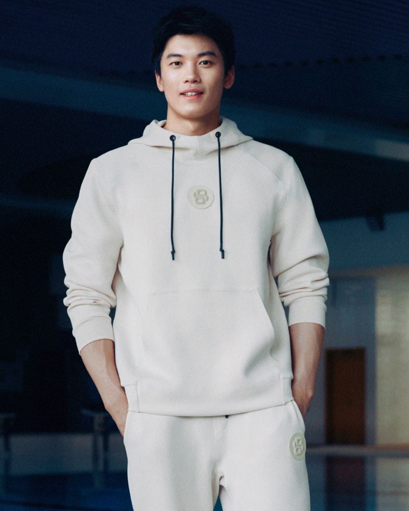 Adult male wearing a light beige hooded sweatshirt and matching sweatpants poses in front of a blurred background.