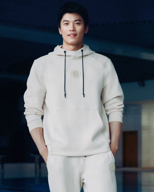 Adult male wearing a light beige hooded sweatshirt and matching sweatpants poses in front of a blurred background.