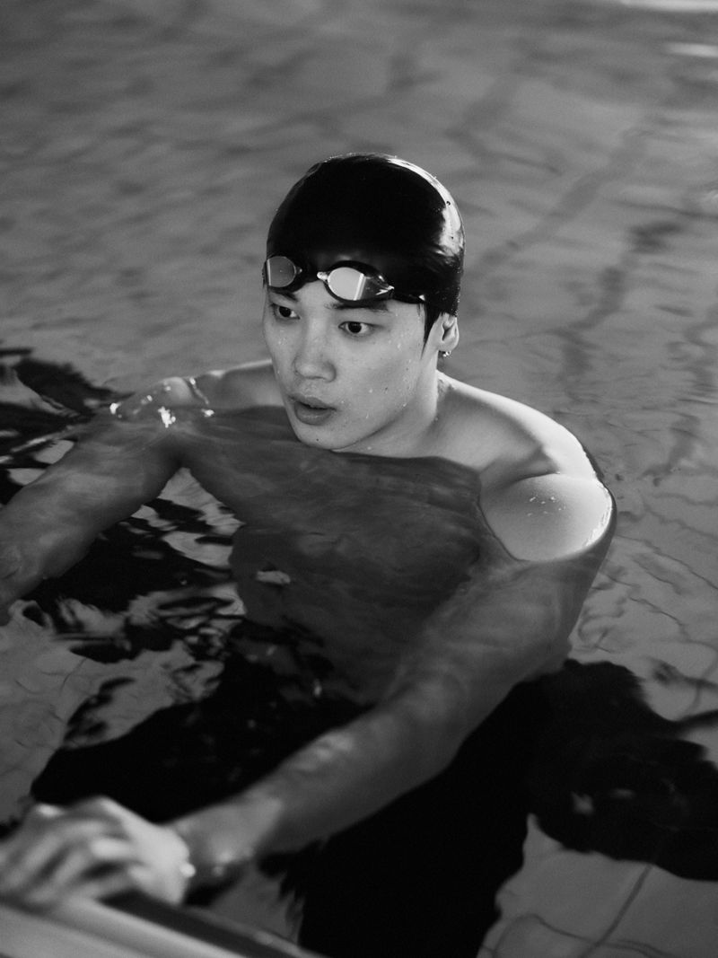 Black and white image of an Asian swimmer in a pool with a swimming cap and goggles, holding onto the side of the pool.