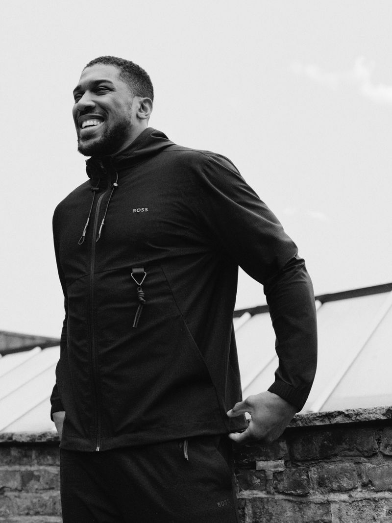 Portrait of smiling adult male in black athletic wear outdoors against a bright sky and building. Features BOSS logo on jacket.