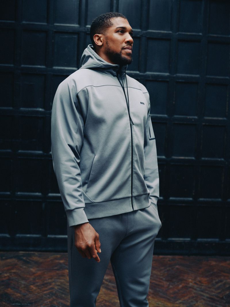 Man in grey hooded tracksuit poses against a dark door. Athletic wear, male fashion, indoor portrait, medium shot.