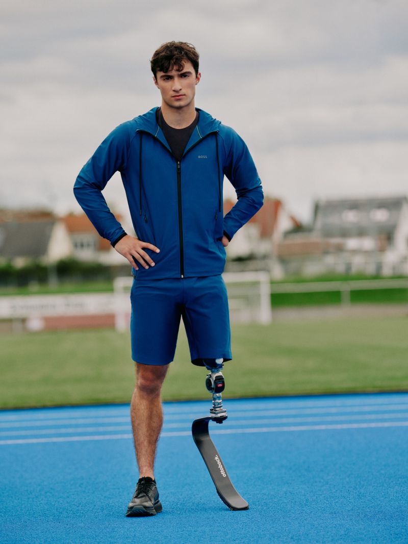 Adaptive athlete stands on blue track in blue athletic suit and black athletic shoes looking towards the camera with grey skies.