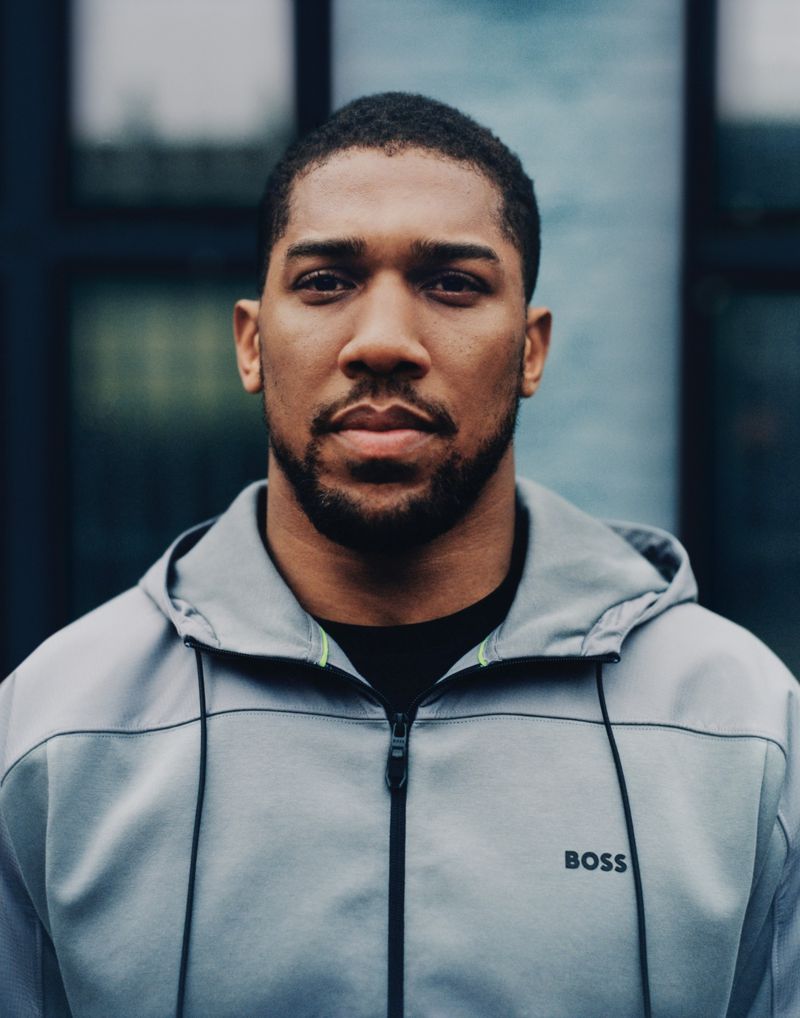 Portrait of an adult male with a beard, wearing a gray hoodie with the BOSS logo, posing outdoors against a building with glass windows.