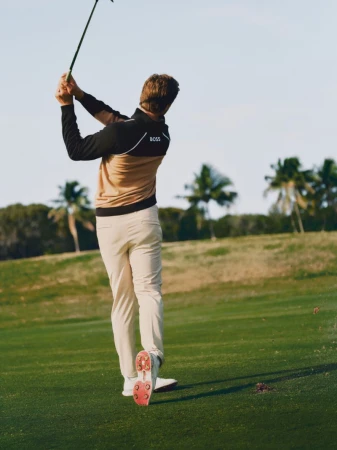 A man wearing beige pants and a brown and black sweater swings a golf club on a green golf course with palm trees in the background.