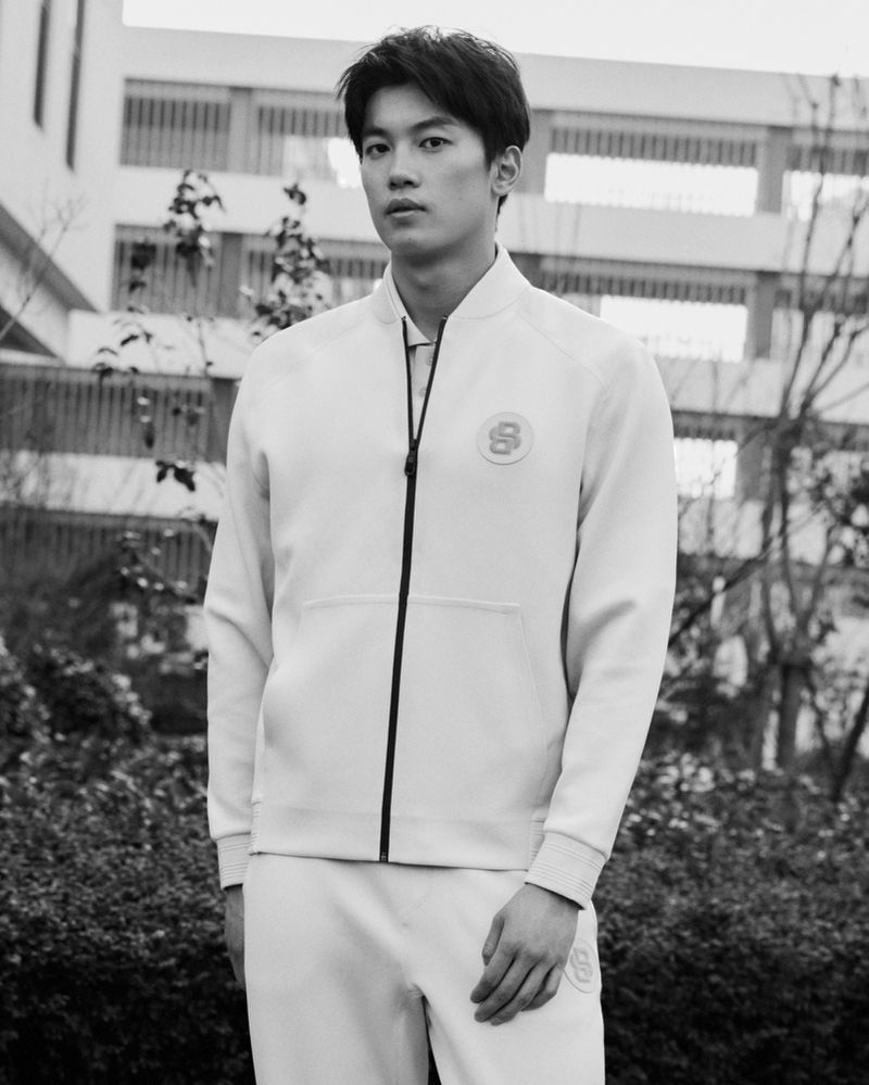 Portrait of an Asian man in a light-colored jacket and pants standing outdoors against a building with trees in a black and white image.