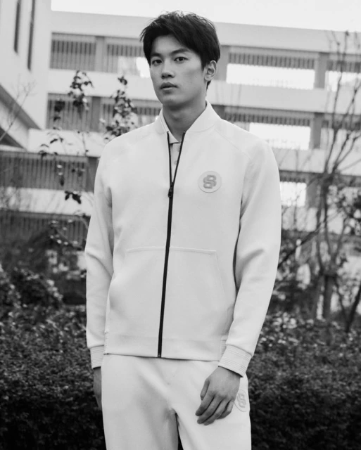Portrait of an Asian man in a light-colored jacket and pants standing outdoors against a building with trees in a black and white image.