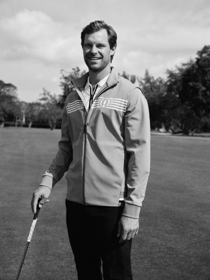 Outdoor portrait of a smiling adult male wearing a jacket and holding a golf club on a sunny day with grass and trees in the background.