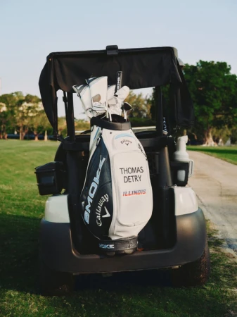 Golf clubs in a golf bag on the back of a golf cart parked on a golf course on a sunny day, trees and green grass visible.