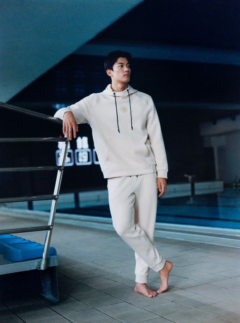 Athletic Asian man in a hoodie and sweatpants leans on a pool ladder indoors, showcasing a blend of relaxation and sporty fashion.