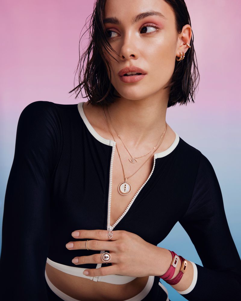 Attractive young woman with fair skin poses in a studio with layered gold jewelry, a black and white swimsuit, and a pink and blue gradient background.
