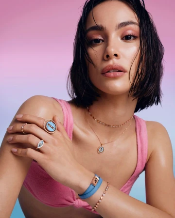 A young woman poses indoors wearing jewelry, a pink top and minimal makeup against a split pink and blue background.