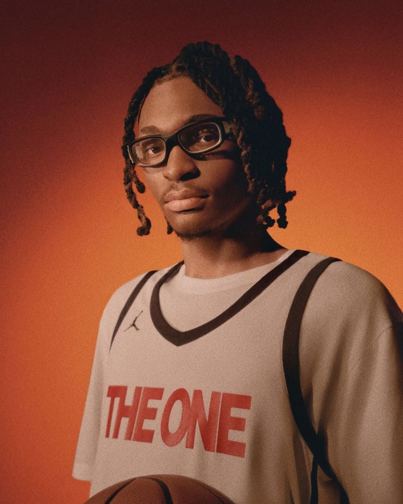 Portrait of a Black male athlete with glasses and dreadlocks in a basketball jersey, holding a ball against an orange gradient background.