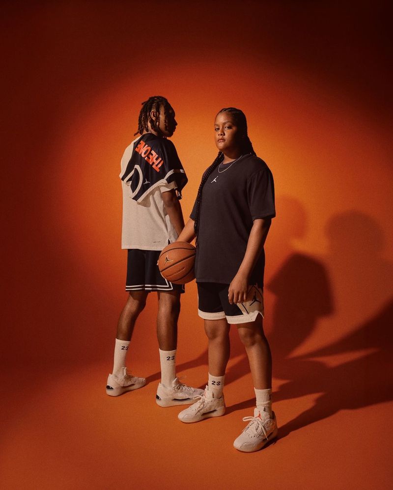 Two athletes in athletic wear pose against an orange background, one holding a basketball and both wearing matching sneakers and sport socks.