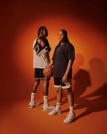 Two athletes in athletic wear pose against an orange background, one holding a basketball and both wearing matching sneakers and sport socks.