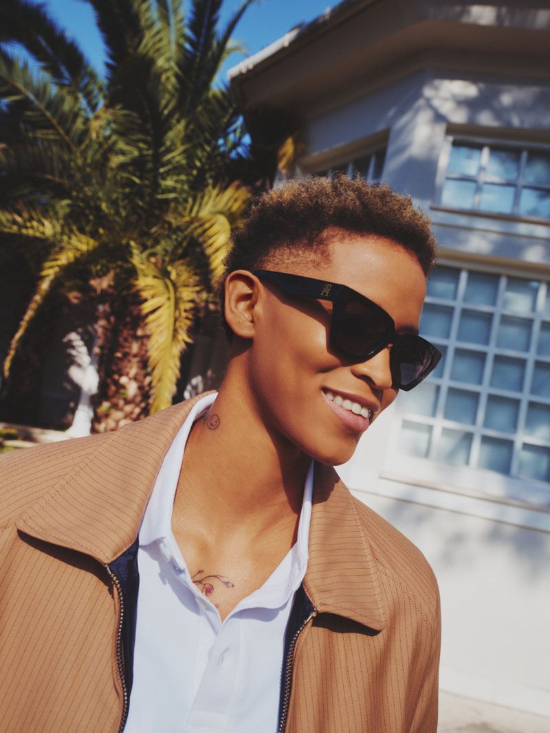 Model wearing dark sunglasses, white polo shirt, and camel jacket outdoors near a palm tree and building exterior.