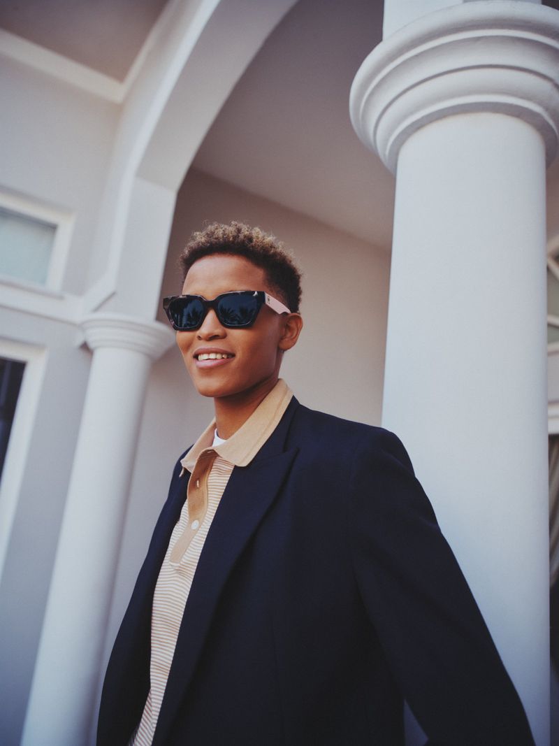 Stylish adult in sunglasses and a blazer smiles outdoors, framed by white architectural columns in a fashion-forward setting.