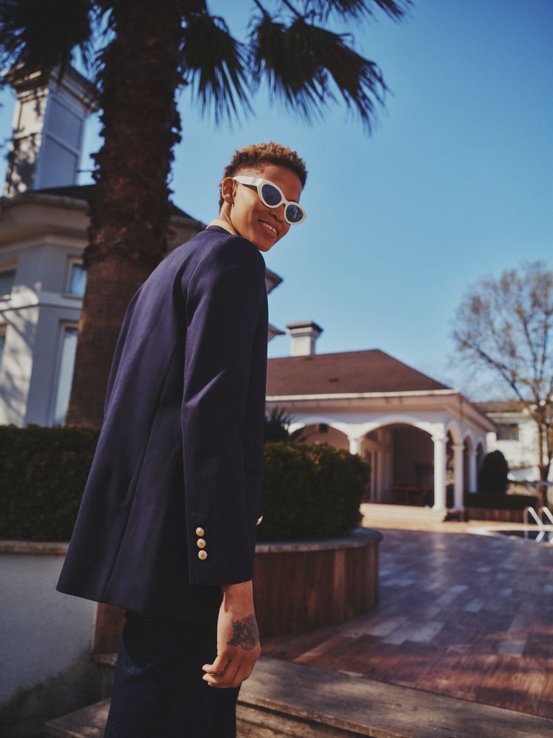 Smiling adult with sunglasses and blue blazer poses on a patio near a house with a red roof and a palm tree against a blue sky.