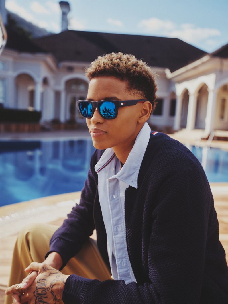 Stylish young adult sits by a pool wearing sunglasses that reflect palm trees and a luxury estate in the background on a sunny day.