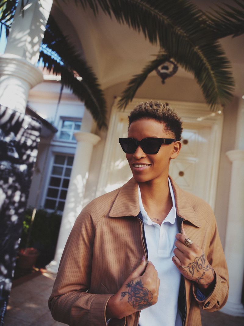 Stylish young adult with sunglasses poses outdoors in a beige jacket and white polo shirt against a sunny building backdrop.