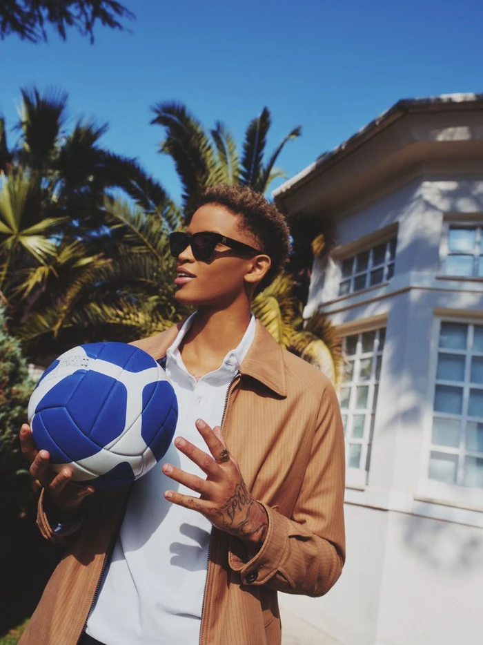 Model wearing dark sunglasses and camel jacket holding a blue and white ball outdoors with palm trees and a building in the background.