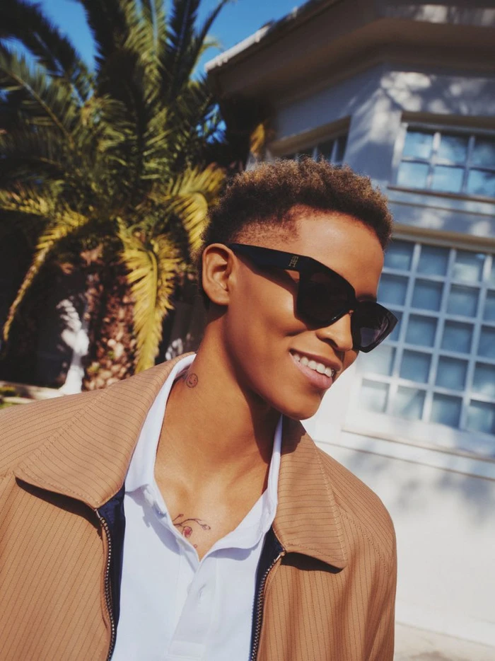 Model wearing dark sunglasses, white polo shirt, and camel jacket outdoors near a palm tree and building exterior.