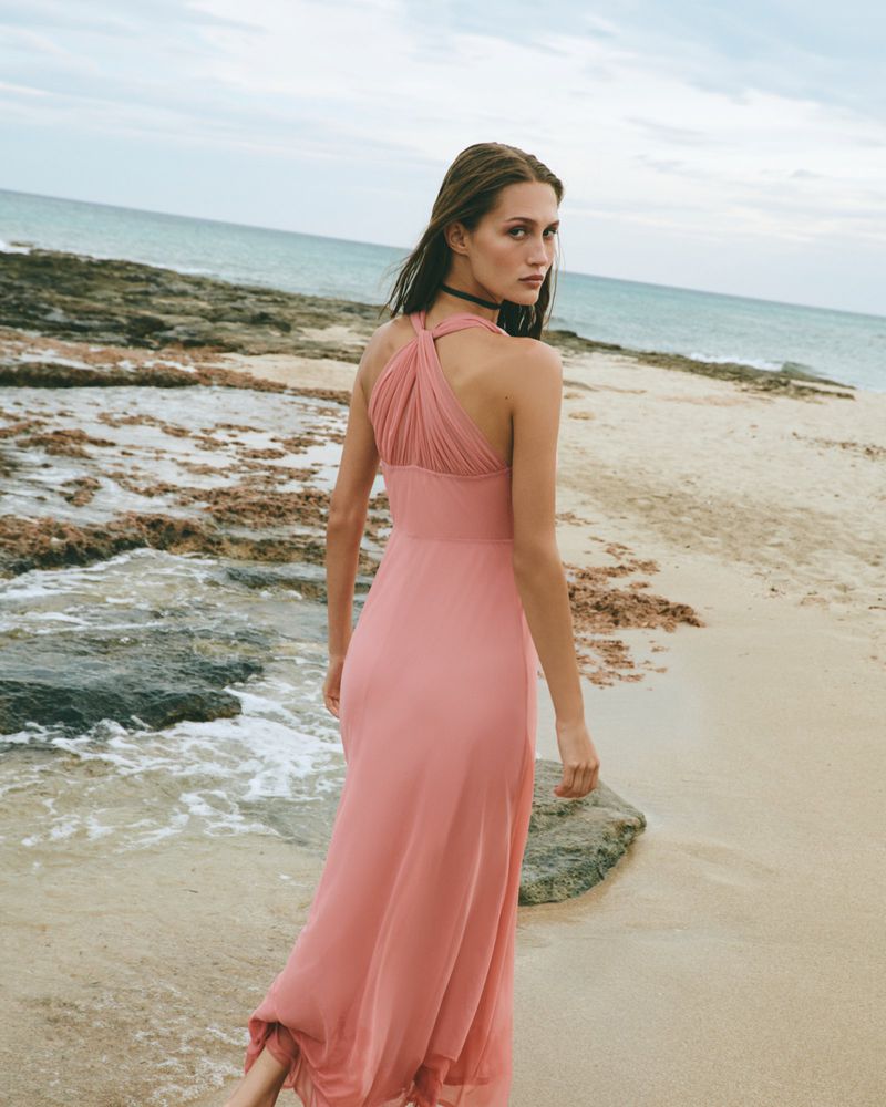 Woman in a long pink halter neck dress walking along a sandy beach near rocky shores and ocean waves under an overcast sky.