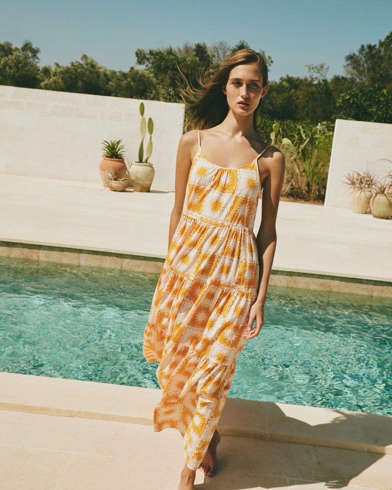 A woman in a yellow-and-white patterned sundress stands near a pool on a sunny day with trees and vegetation in the background.