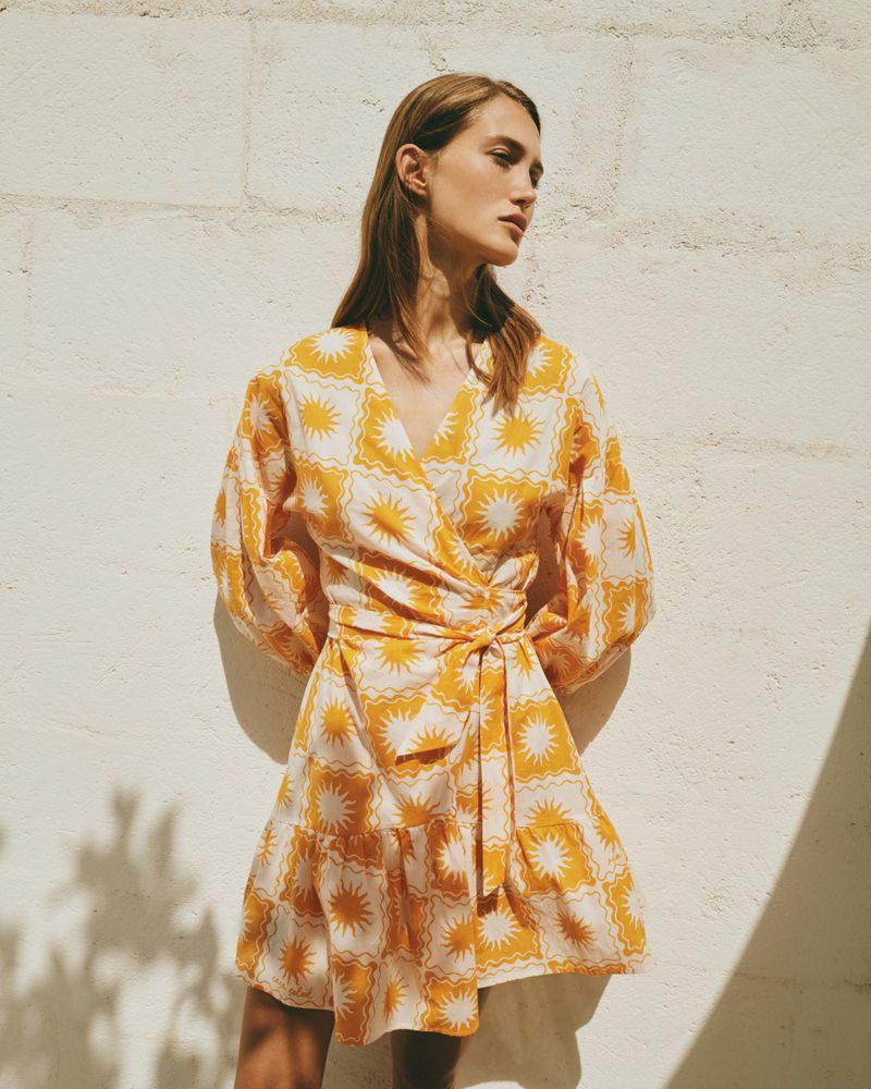 A fair-skinned woman poses in a short-sleeved, yellow-and-white patterned wrap dress, outdoors against a textured wall with shadows.