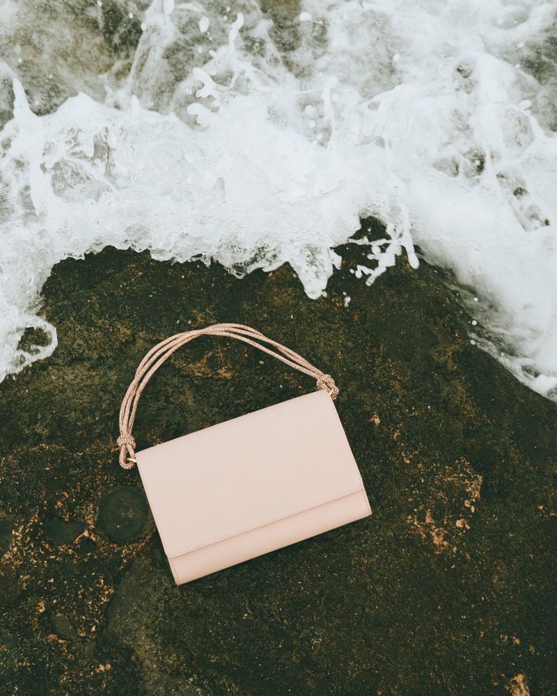 Beige purse with rope straps laying on a dark rock with white sea foam in the background, shot from a high angle in natural light.