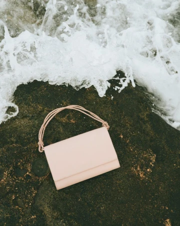 Beige purse with rope straps laying on a dark rock with white sea foam in the background, shot from a high angle in natural light.