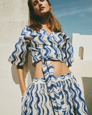 Young woman in wavy-patterned linen crop top and skirt, posing against a textured wall with blue sky, summer fashion, outdoor setting.