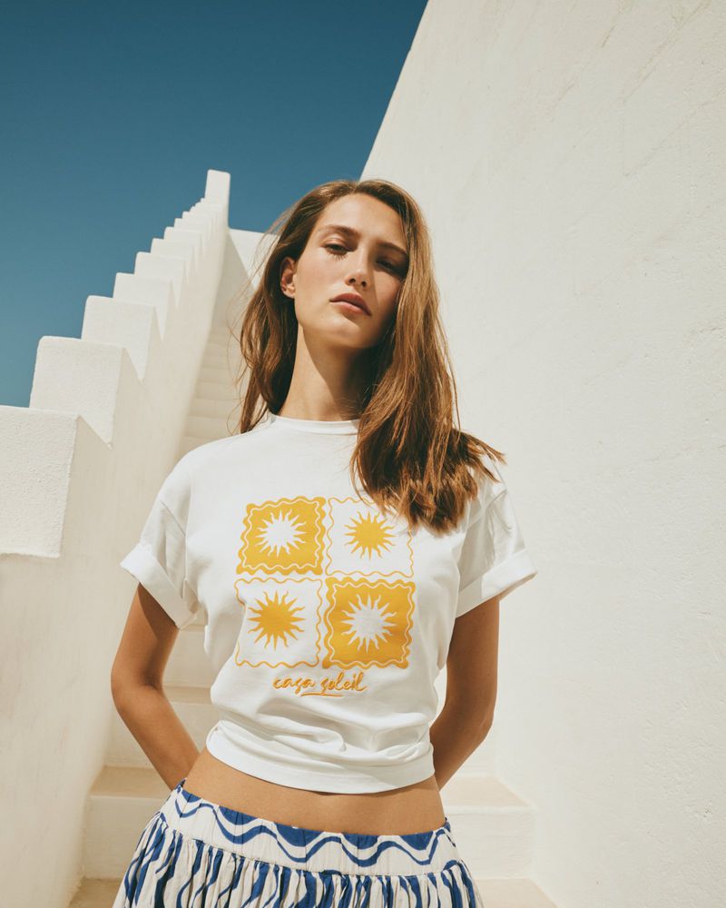 Woman in white tee with sun graphics and patterned skirt poses on steps against white wall and blue sky, natural light.
