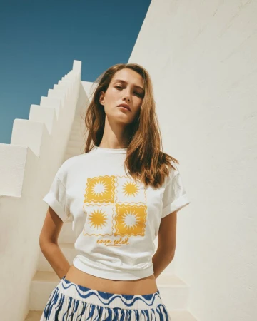 Woman wearing graphic white t-shirt and blue patterned skirt standing before white stairs under blue sky