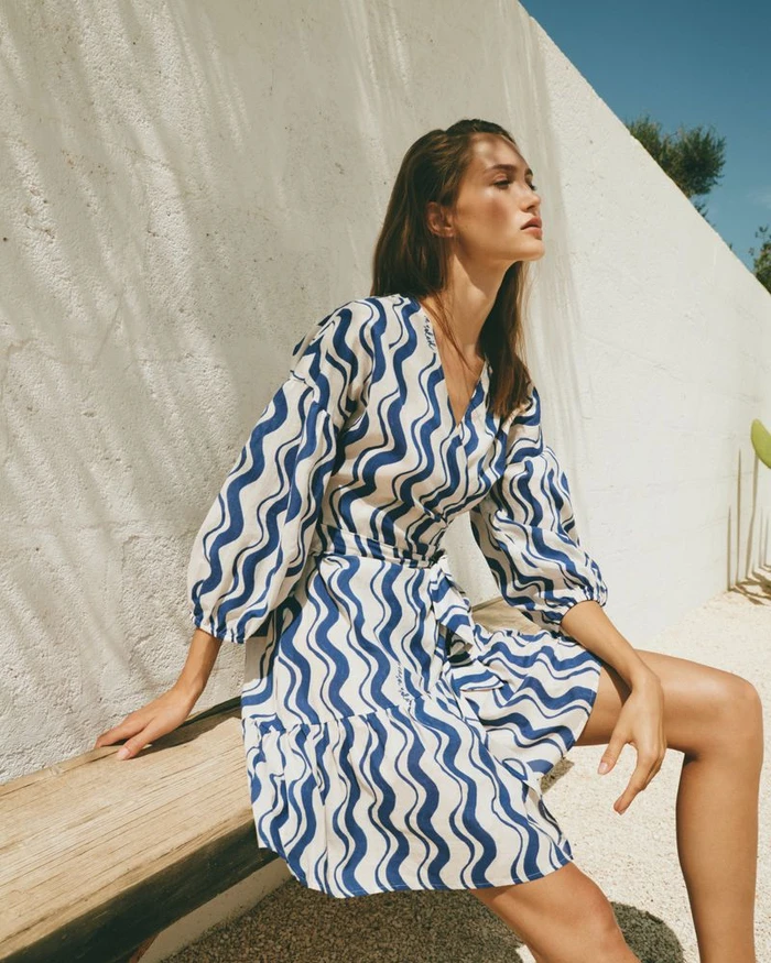 Model wearing a blue and white patterned wrap dress seated on a wooden bench against a white exterior wall.