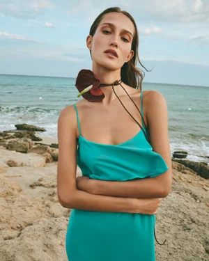 Model in a bright turquoise slip dress wearing a dark anthurium flower choker on a rocky beach by the ocean.